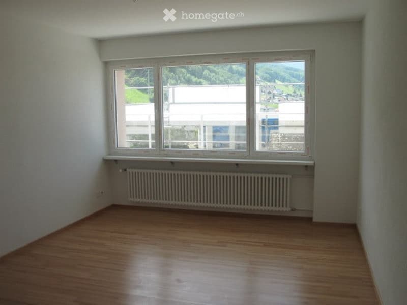Empty room with wooden flooring, white walls, and a large window offering a view of the outside. A radiator is visible below the window.