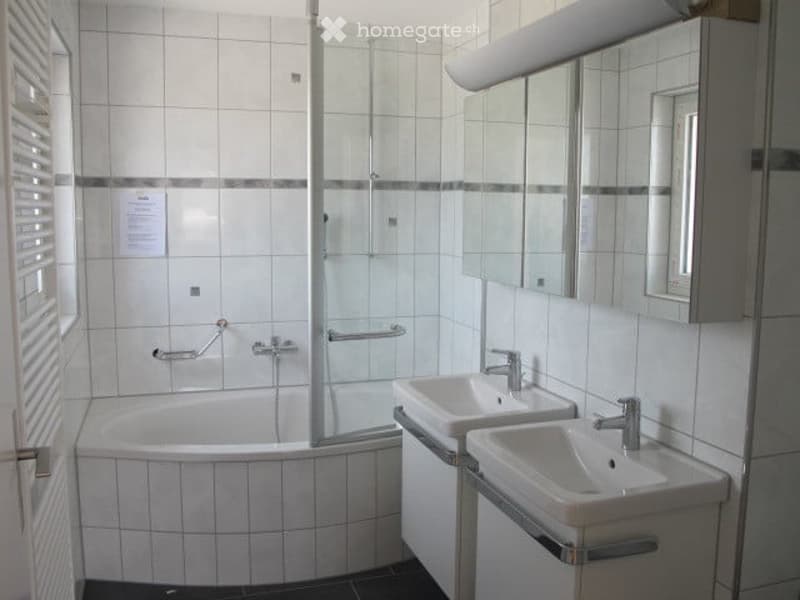 Modern bathroom with white tiled walls and floor, featuring a bathtub, two sinks, a mirror, and a glass-enclosed shower area.