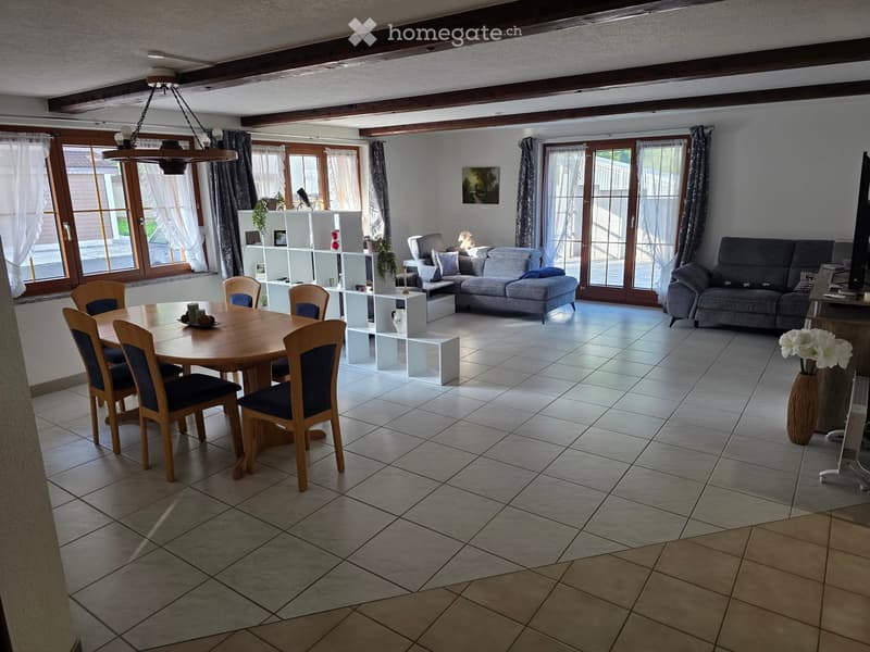 Bright living and dining area with tiled flooring, large windows, and wooden furniture including a sofa, dining table, and chairs.