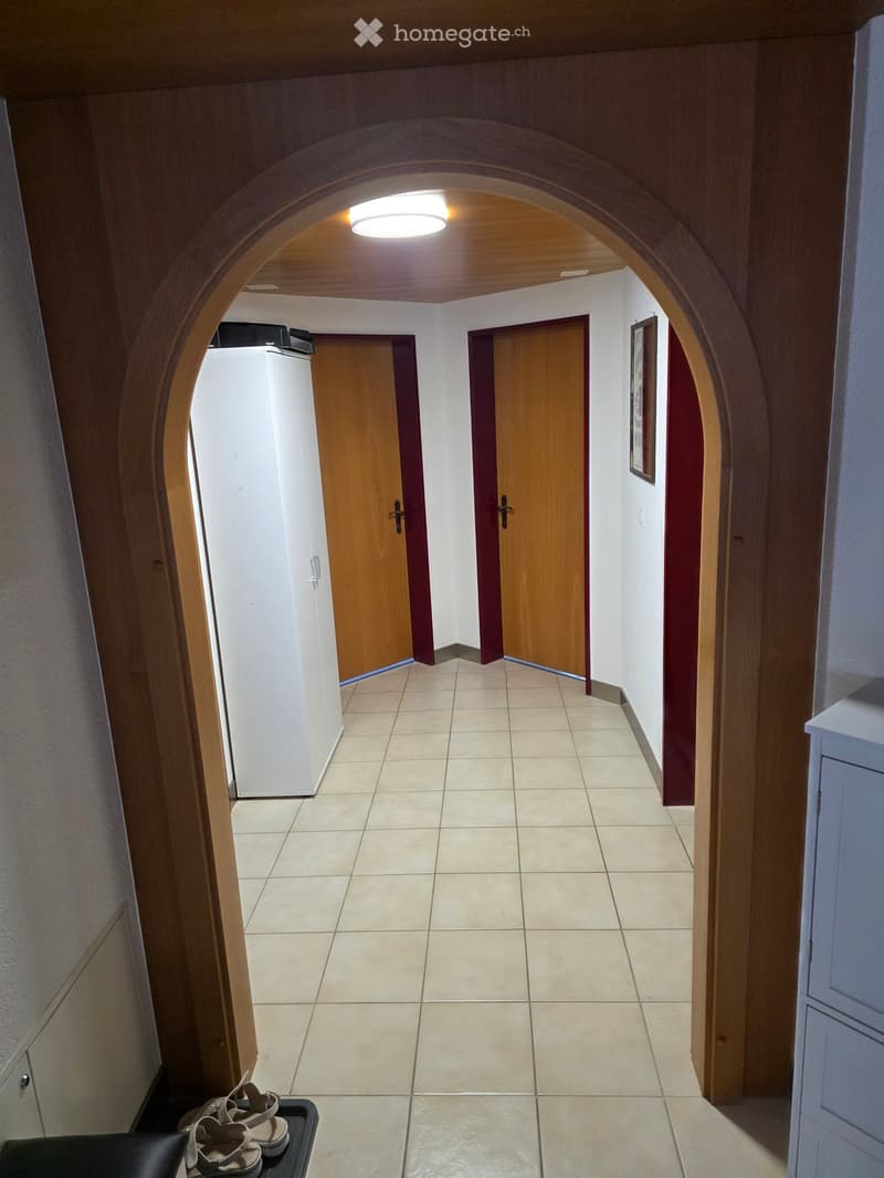 A well-lit hallway with beige tiled flooring, wooden doors, and a curved ceiling with a central light fixture. The walls are painted white with wooden paneling on one side.