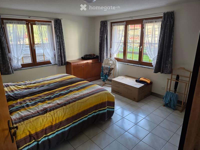 Bedroom with a double bed, wardrobe, chair, and a box on the floor. The room has large windows with curtains and a radiator below them.