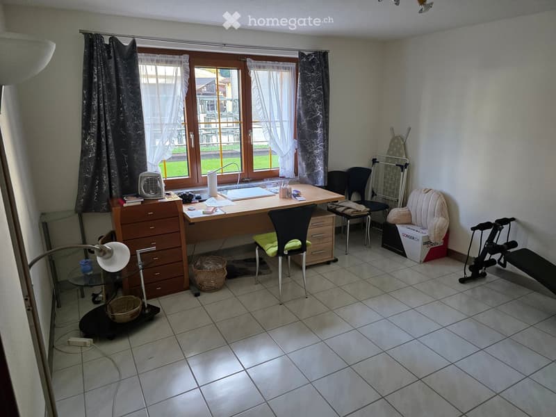 A furnished room with a desk, chair, sofa, lamp, fan, drawer unit, wicker basket, exercise bike, ironing board, and other items.