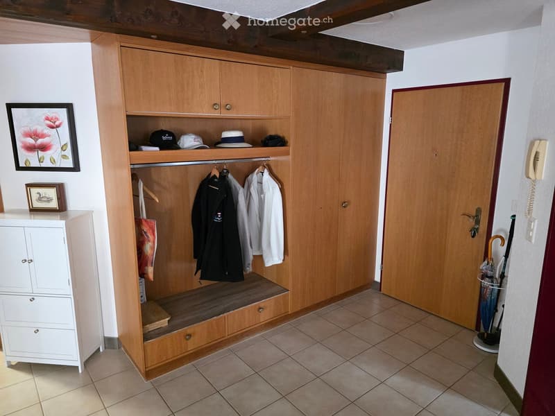 A well-organized hallway with a wooden coat rack, a small bench, and a white cabinet. The coat rack holds jackets, hats, and other items, while the bench provides a place to sit. A framed picture and a small table with a decorative item are also visible.