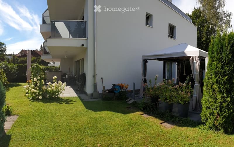 Well-maintained garden with a lawn, flower beds, a covered patio area, and a modern house with a balcony and large windows.