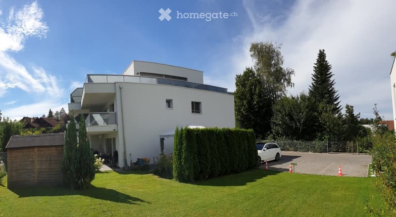 Modern two-story house with white exterior, large windows, and a balcony. The property includes a well-maintained lawn, a wooden shed, and a parking area with a white car.