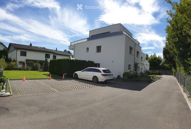 Modern two-story house with white exterior, flat roof, and balcony on the upper level. A white SUV is parked on the driveway, and there is a well-maintained lawn and hedge in the front yard.