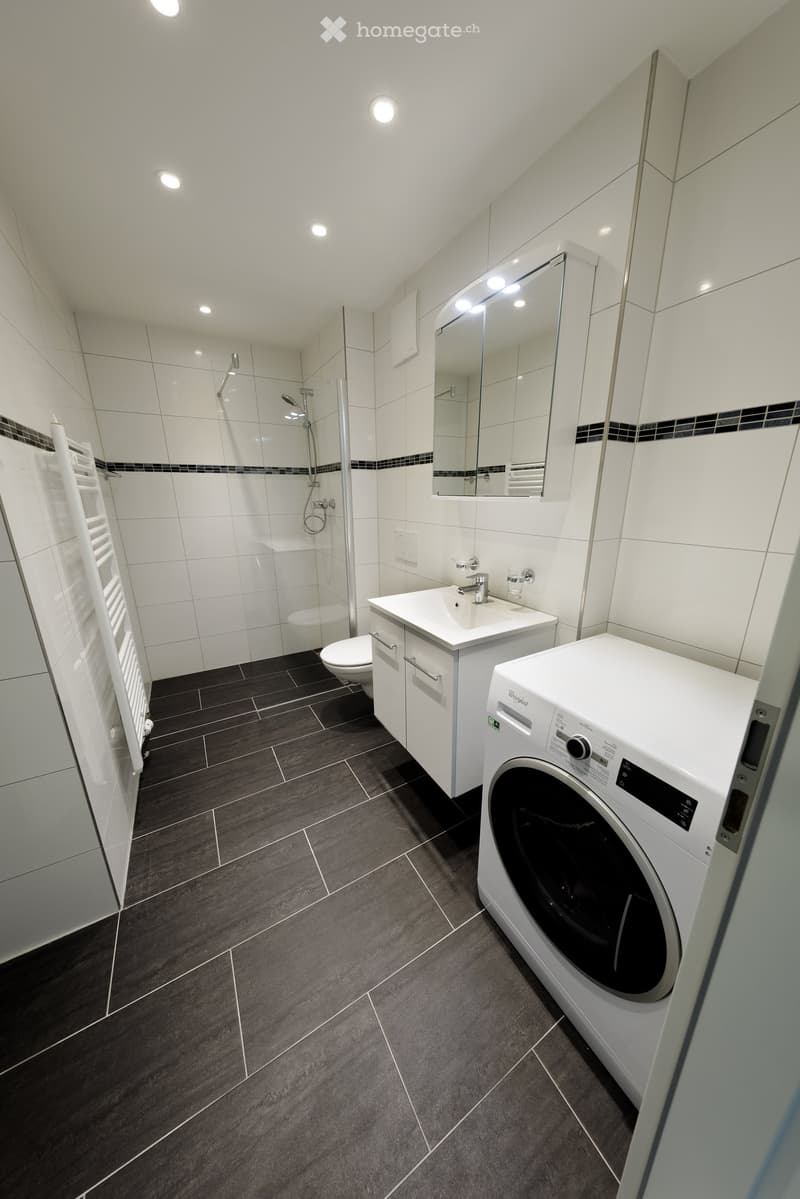 Modern bathroom with white tiles, dark grey floor, sink, mirror, toilet, shower, and washing machine.
