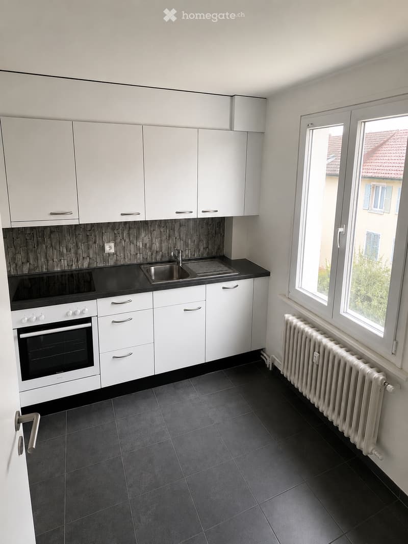 Modern kitchen with white cabinets, black countertop, stainless steel sink, and built-in oven. Dark tiled floor and radiator visible.