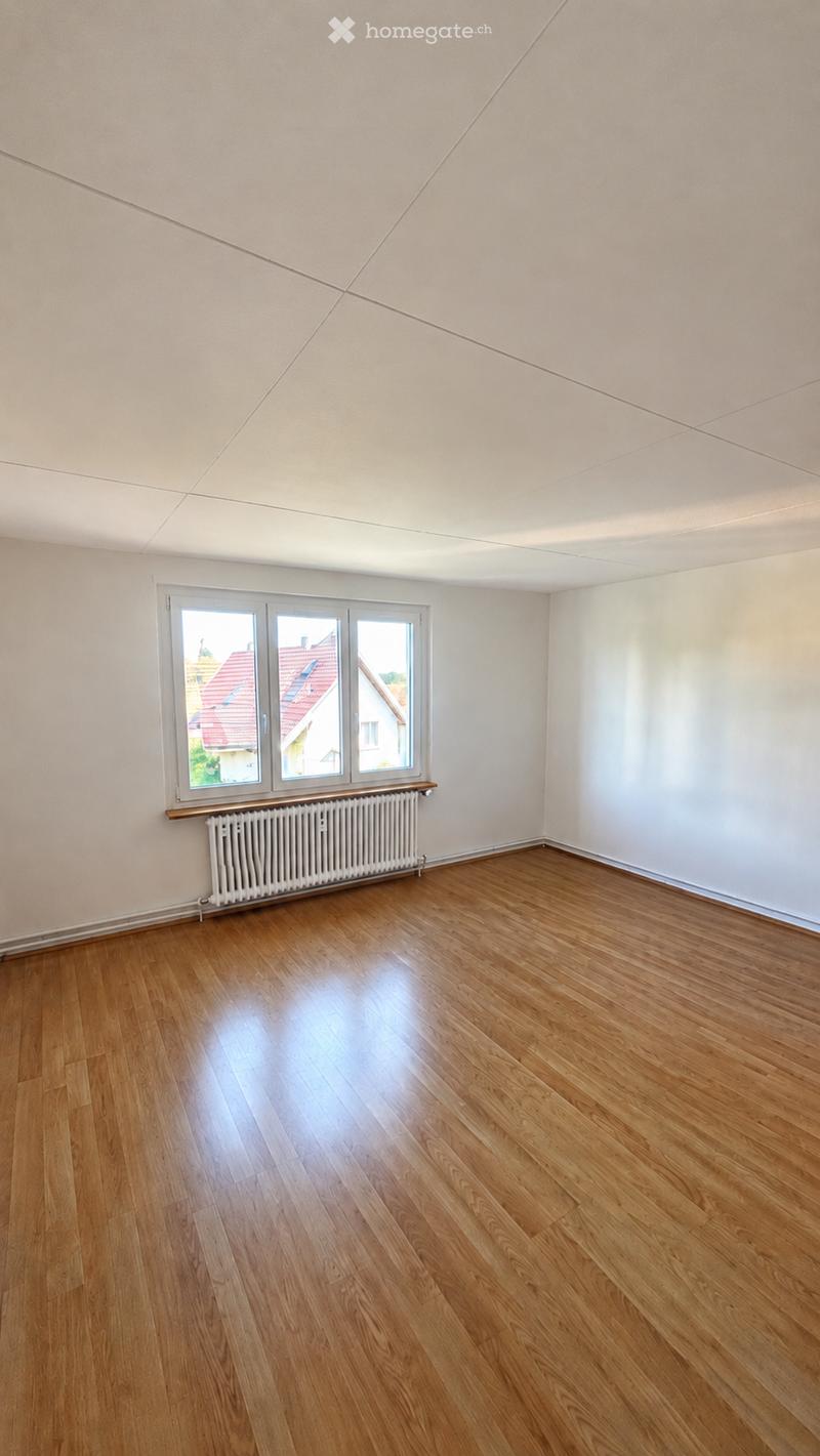 Empty room with wooden flooring, white walls, and large windows offering a view of neighboring houses.