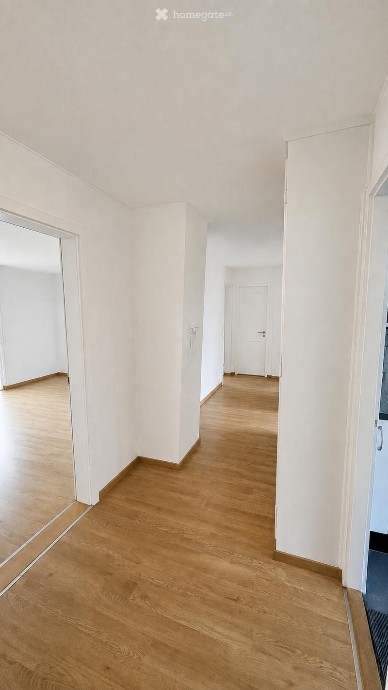 Empty hallway with light wooden flooring and white walls, featuring several doors and a large mirror on one side.