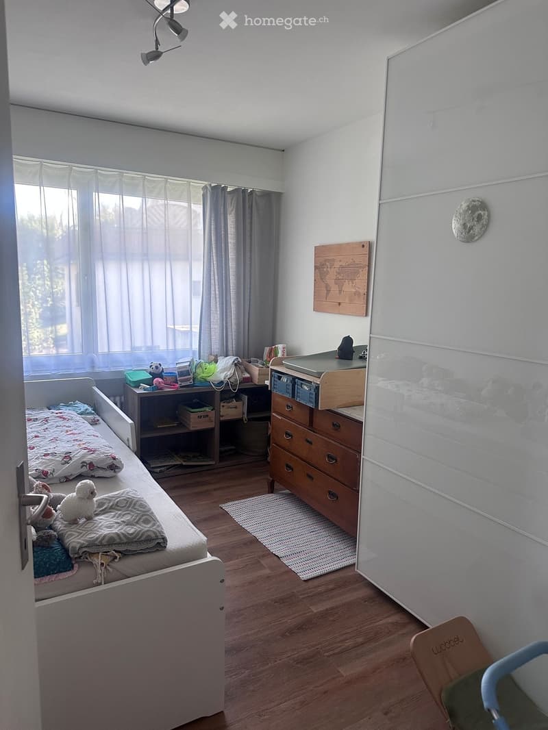 A well-lit bedroom with a single bed, nightstand, drawer unit, and a small table with various items. The room features wooden flooring, a large window with curtains, and decorative elements like a world map and a mirror.