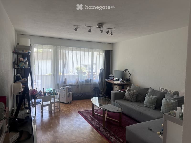 A modern living room with a grey sofa, coffee table, glass coffee table, small dining table, and a laptop on a desk. The room features wooden flooring, large windows with sheer curtains, and a shelf with decorative items.