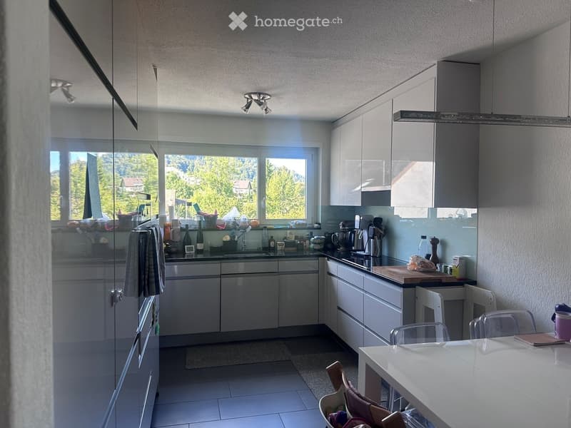 Modern kitchen with white cabinets, stainless steel appliances, and a dining area with a white table and chairs. The kitchen features a large window with a scenic view of greenery and mountains.