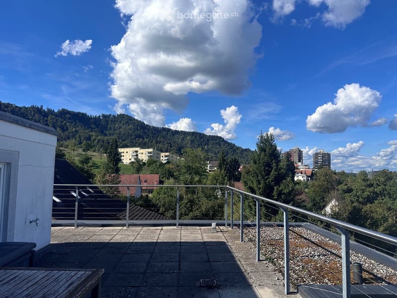 Roof terrace with metal railing, concrete tiles, and a scenic view of a town and hills under a partly cloudy sky.