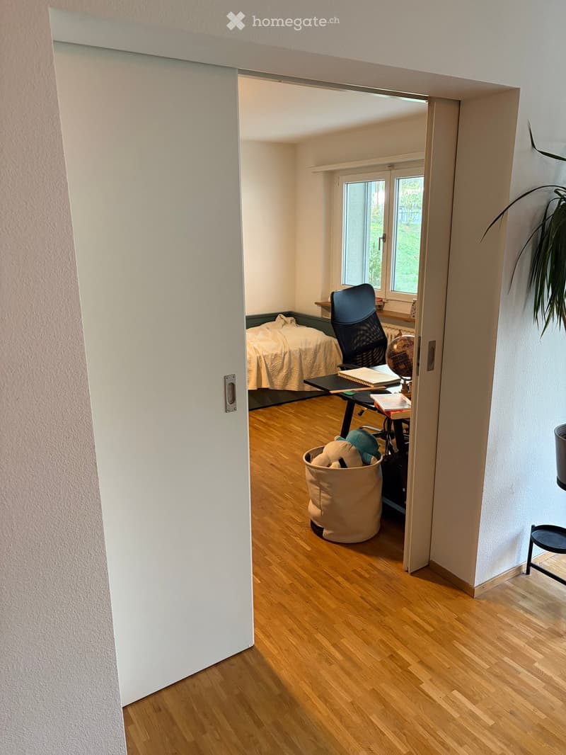 A furnished room with a wooden floor, a desk with a chair, a laptop, books, a globe, a bed, a beige bag, and a small stool.