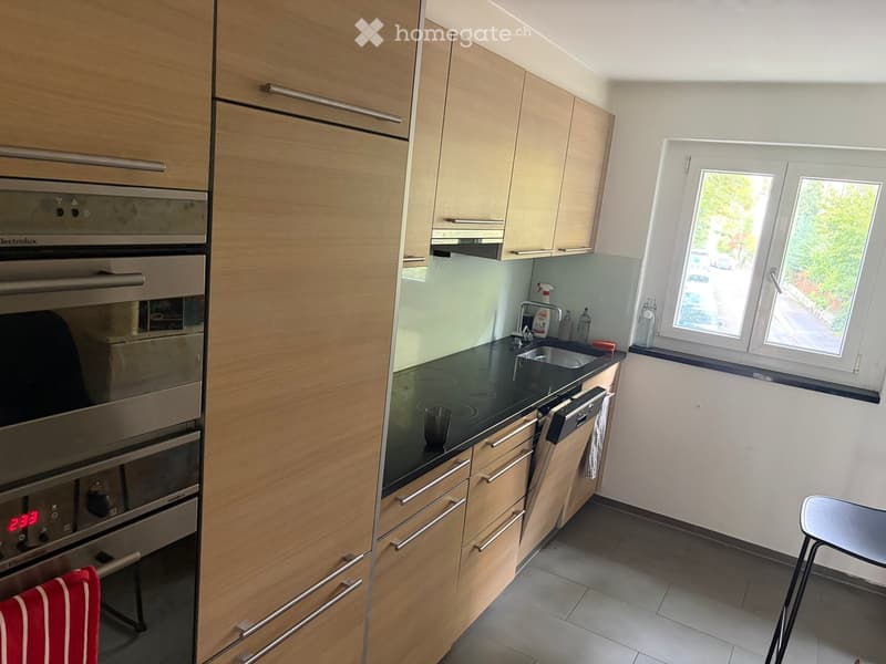 Modern kitchen with light wood cabinets, stainless steel appliances, and a black countertop.