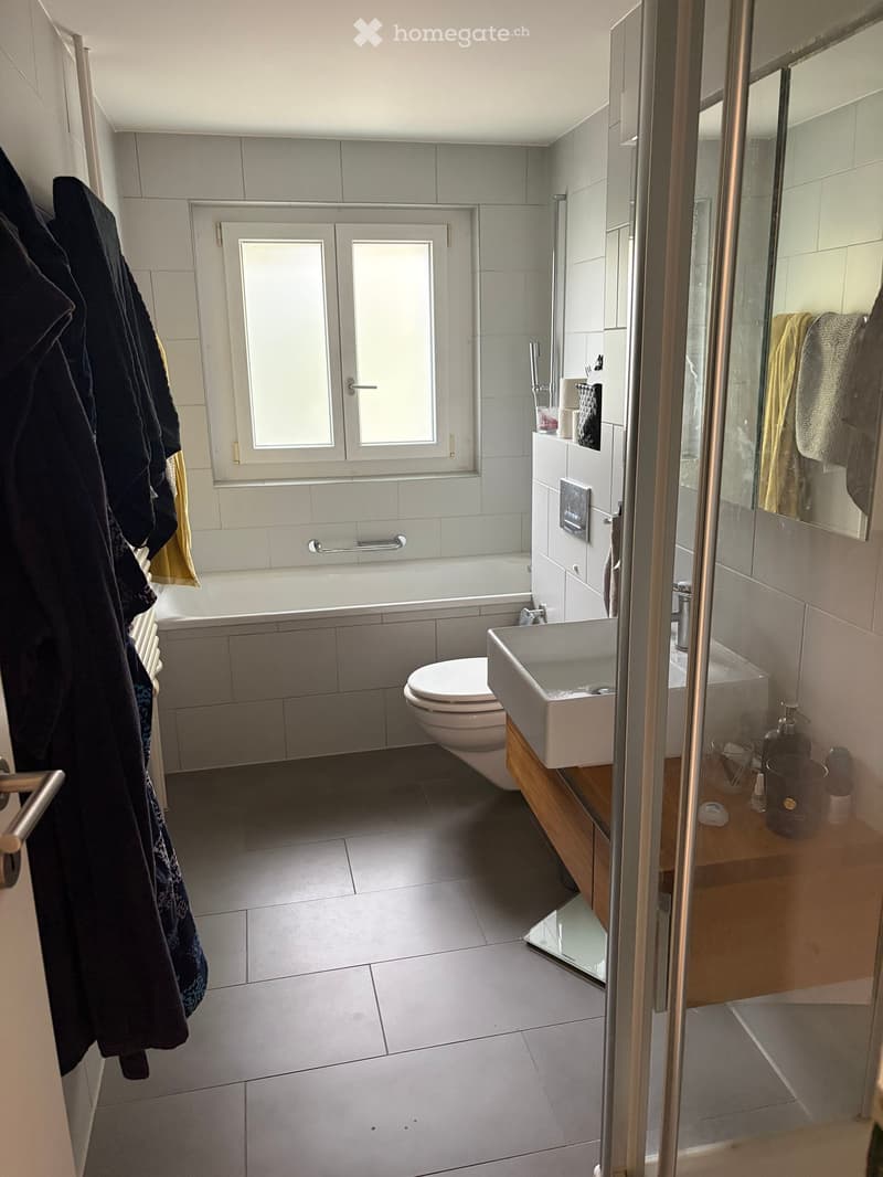 Modern bathroom with a bathtub, toilet, sink, and mirror. Features tiled walls and floor, a window with frosted glass, and a wooden vanity with a sink and various toiletries.