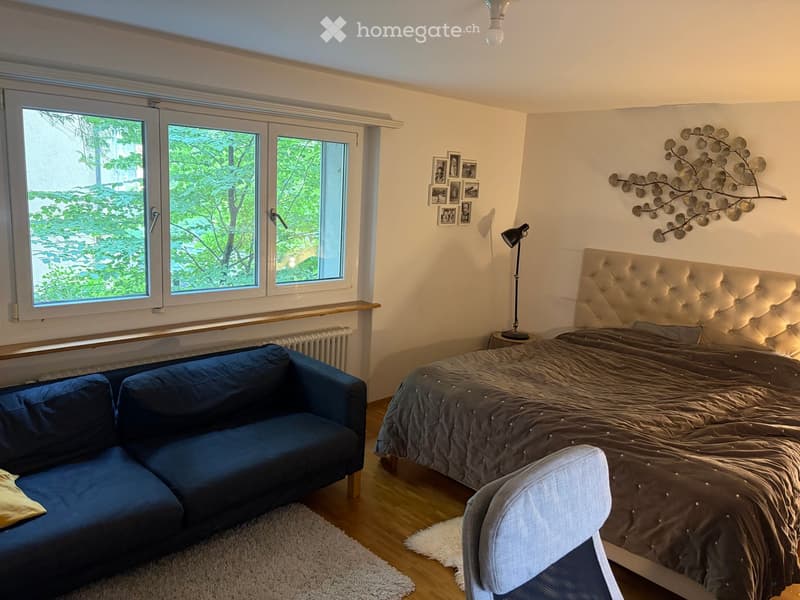 Bright bedroom with large windows, a tufted headboard bed, a blue sofa, a white chair, and wall art.