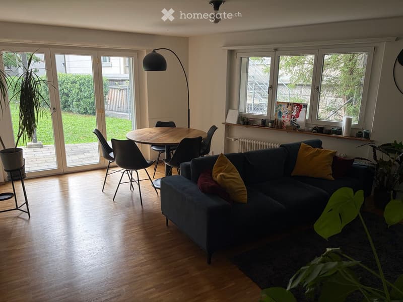 Bright living room with wooden flooring, large windows, and a sliding glass door leading to a patio. Features a dark blue sectional sofa with yellow and red pillows, a round wooden dining table with four black chairs, and a tall floor lamp. Potted plants