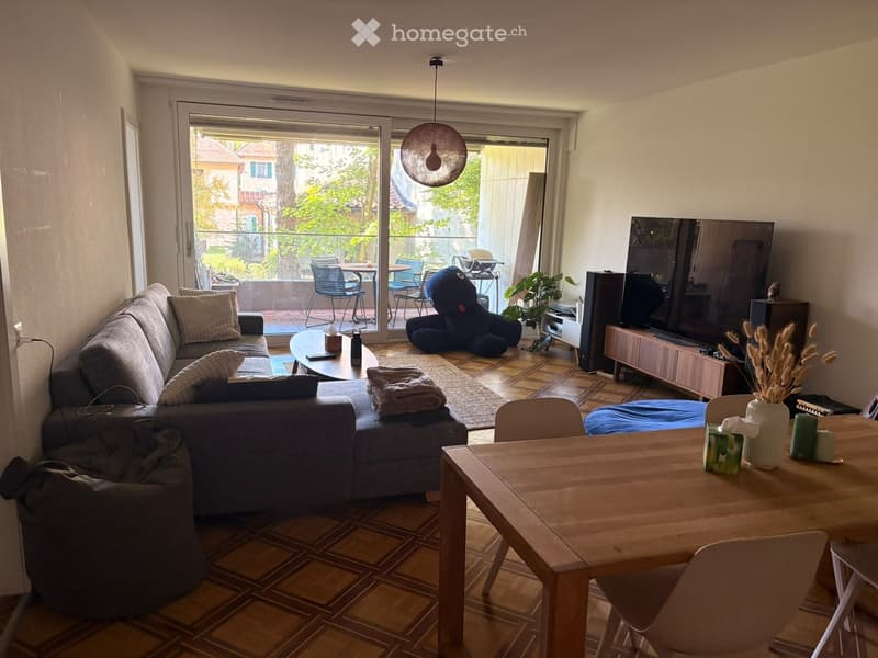 Modern living room with a grey sectional sofa, coffee table, dining table, chairs, TV, and a large window leading to a balcony.