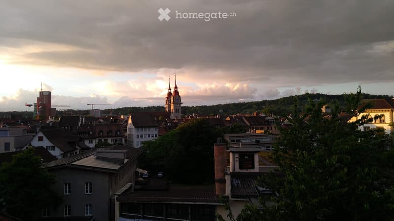 A panoramic view of a cityscape with a prominent church tower, surrounded by residential and commercial buildings under a cloudy sky.