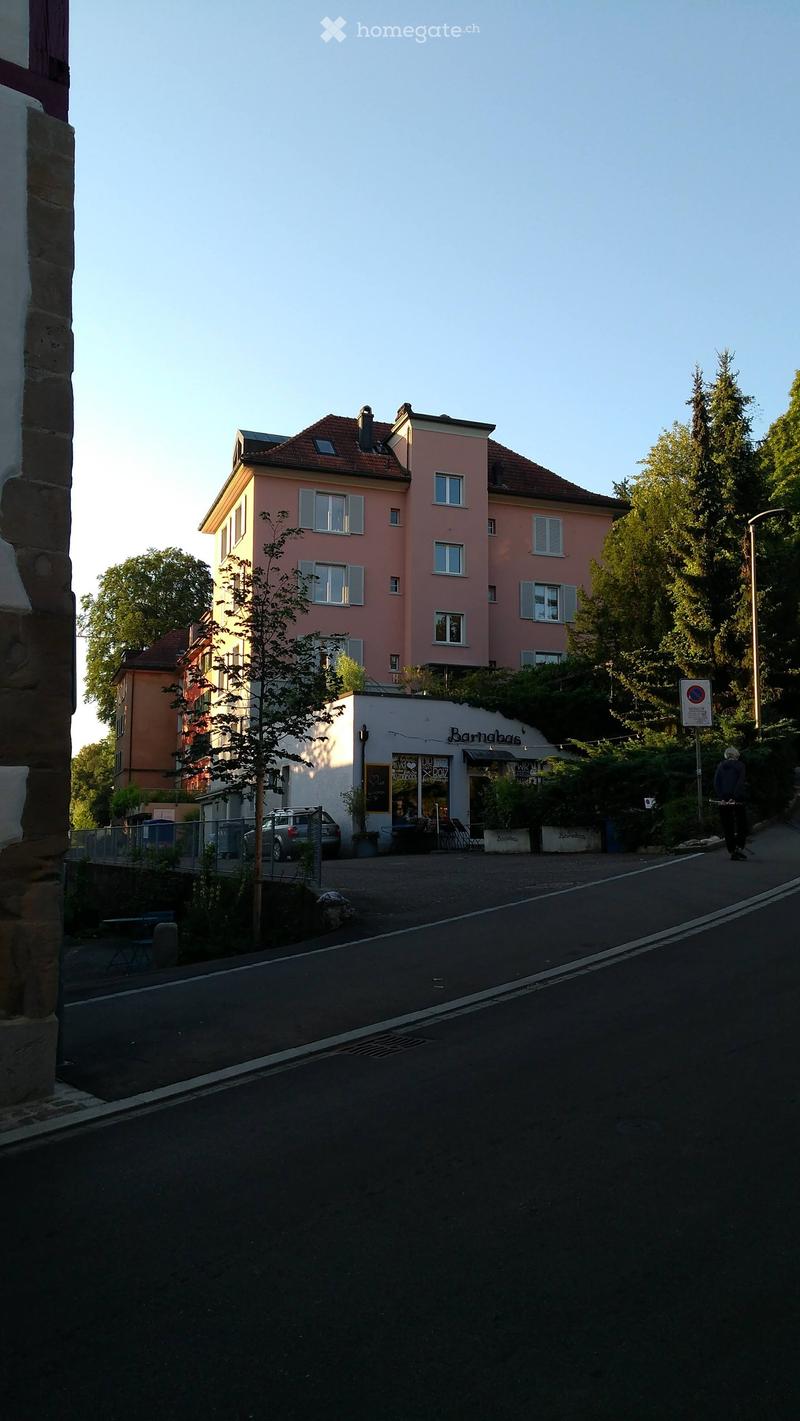 A multi-story residential building with pink facade and multiple windows, adjacent to a small shop with outdoor seating and a car parked nearby.