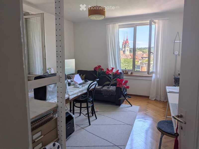 Bright living room with wooden flooring, large windows offering a cityscape view, a sofa, a laptop on a table, and a flower vase on the floor.