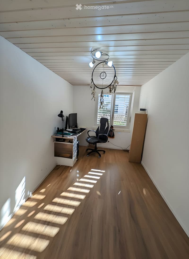 Bright, unfurnished room with wooden flooring, white walls, and a slanted ceiling. Features a desk with a computer, a chair, and a decorative hanging light fixture.
