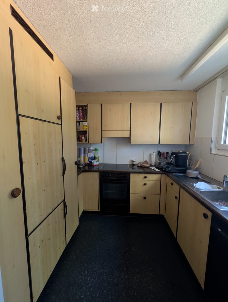 A modern kitchen with light wooden cabinets, a black countertop, and various appliances including an oven, sink, and fridge.