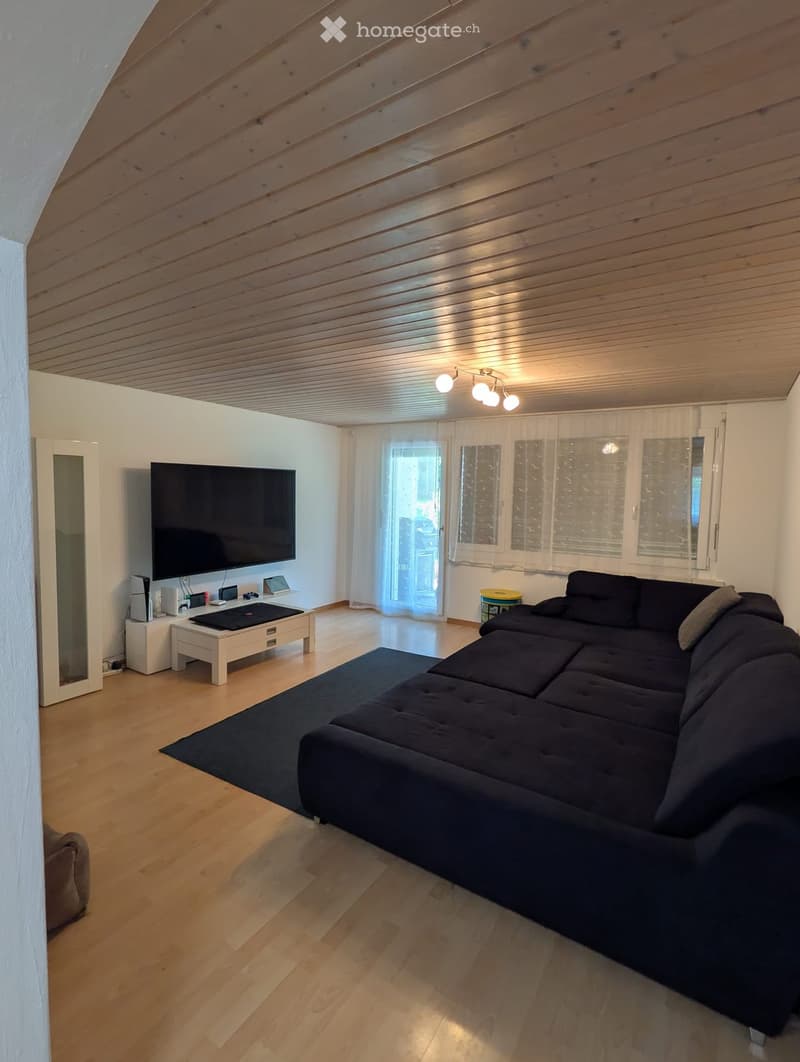 Modern living room with wooden flooring, a large sectional sofa, coffee table, flat-screen TV, and a mirror on the wall.
