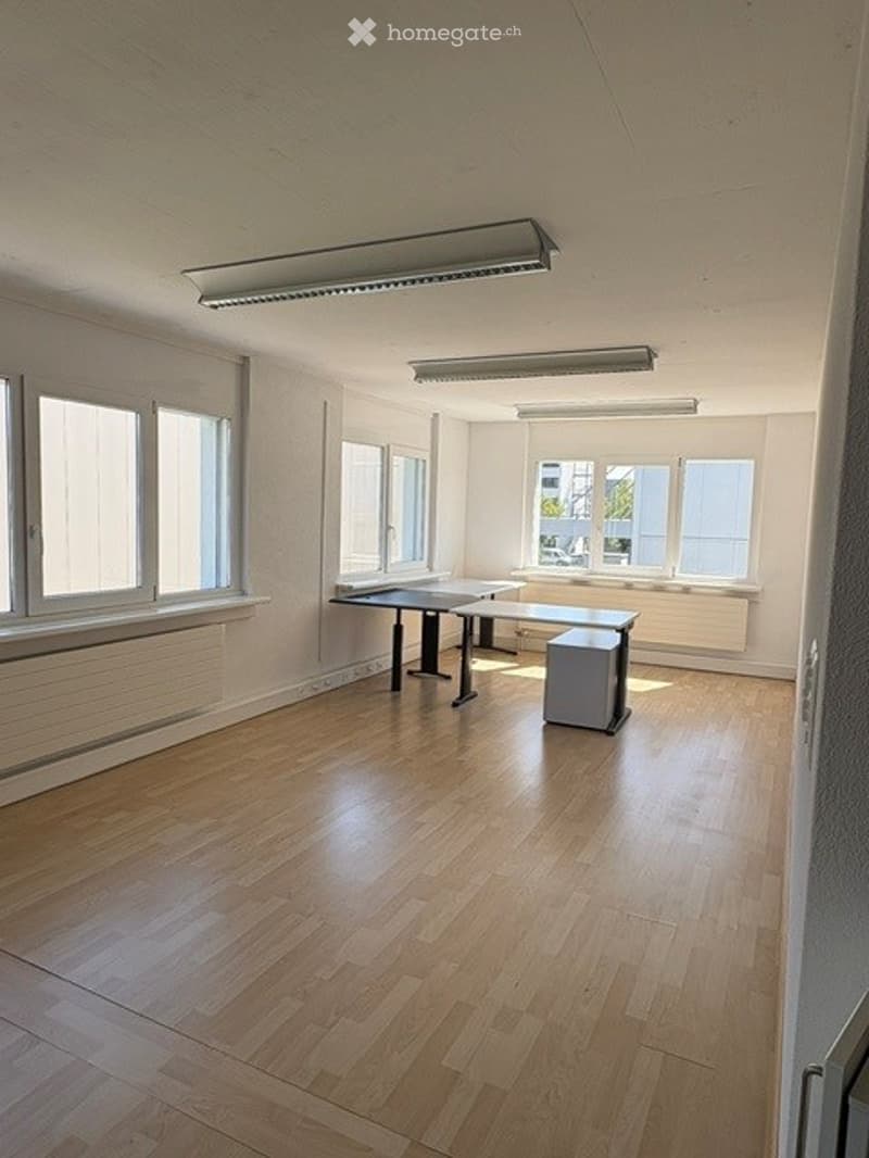Empty room with wooden flooring, large windows, and minimal furniture including a small table and chair.