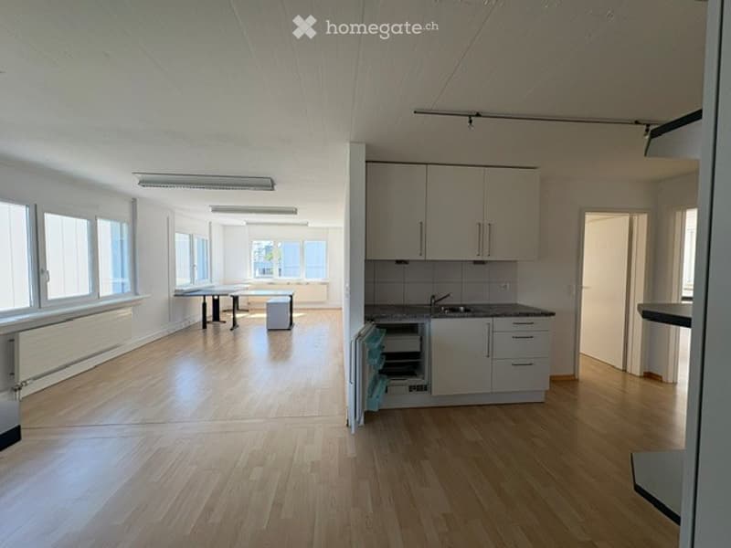 Bright, open-plan space with light wooden flooring, white walls, and large windows. Includes a small table with a stool, a fitted kitchen area with cabinets, a sink, and a faucet.