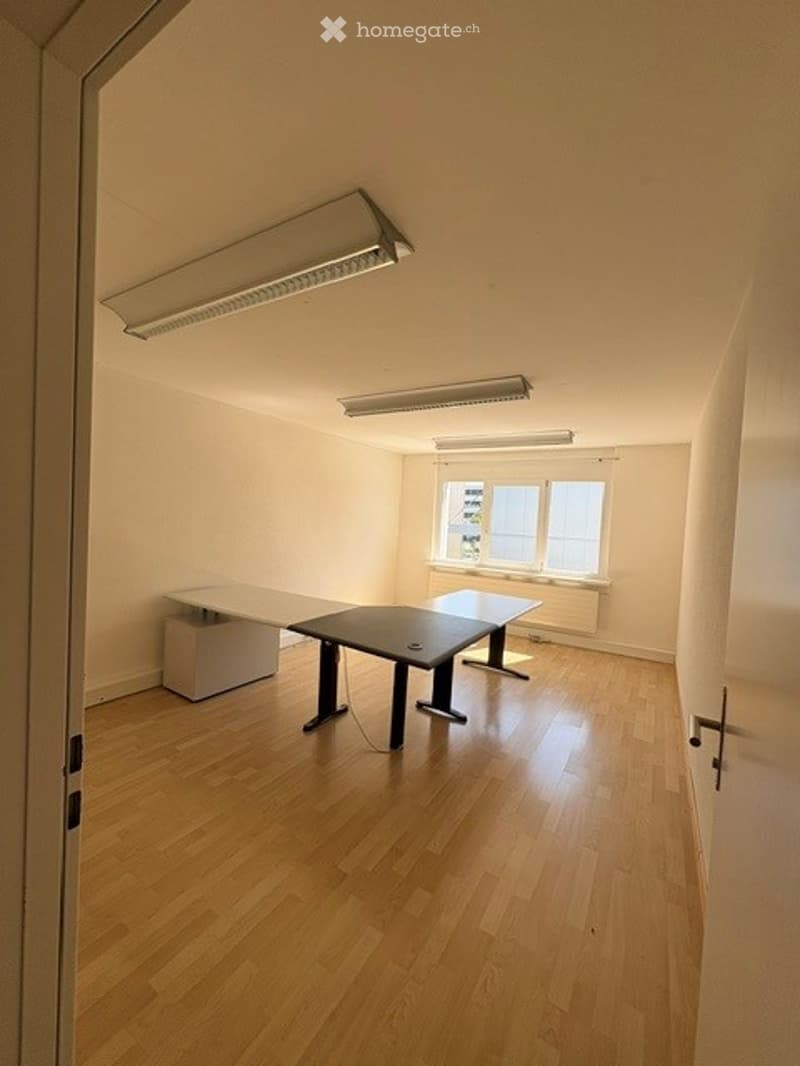 Bright, unfurnished room with wooden flooring, white walls, and large windows. Contains a single table and chair.