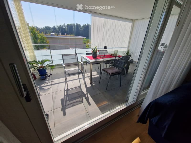 Modern balcony with large glass doors, tiled floor, and outdoor furniture including a table, chairs, and a flower pot.