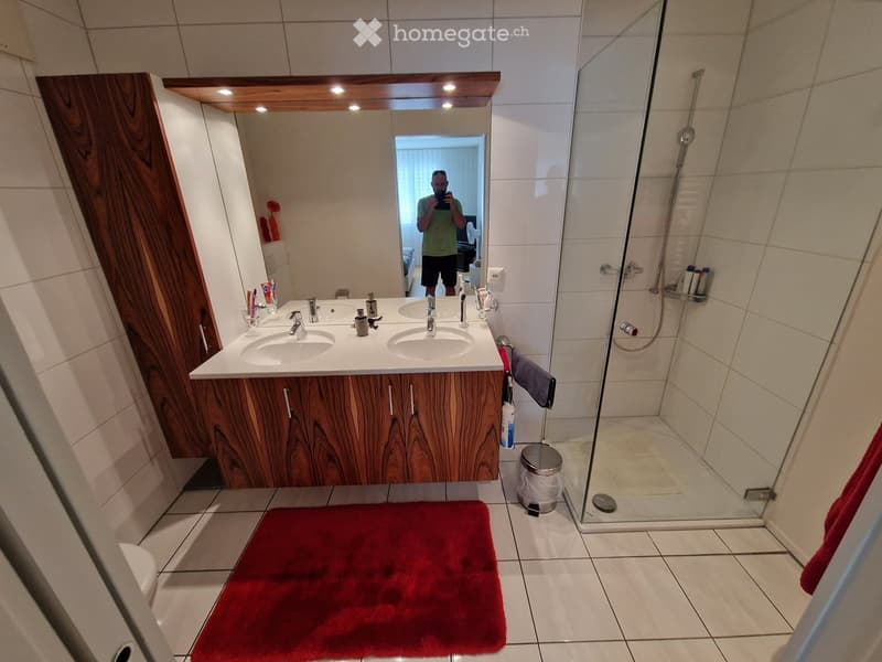 Modern bathroom with double sink vanity, wooden cabinetry, large mirror, glass shower enclosure, and red bath mat.