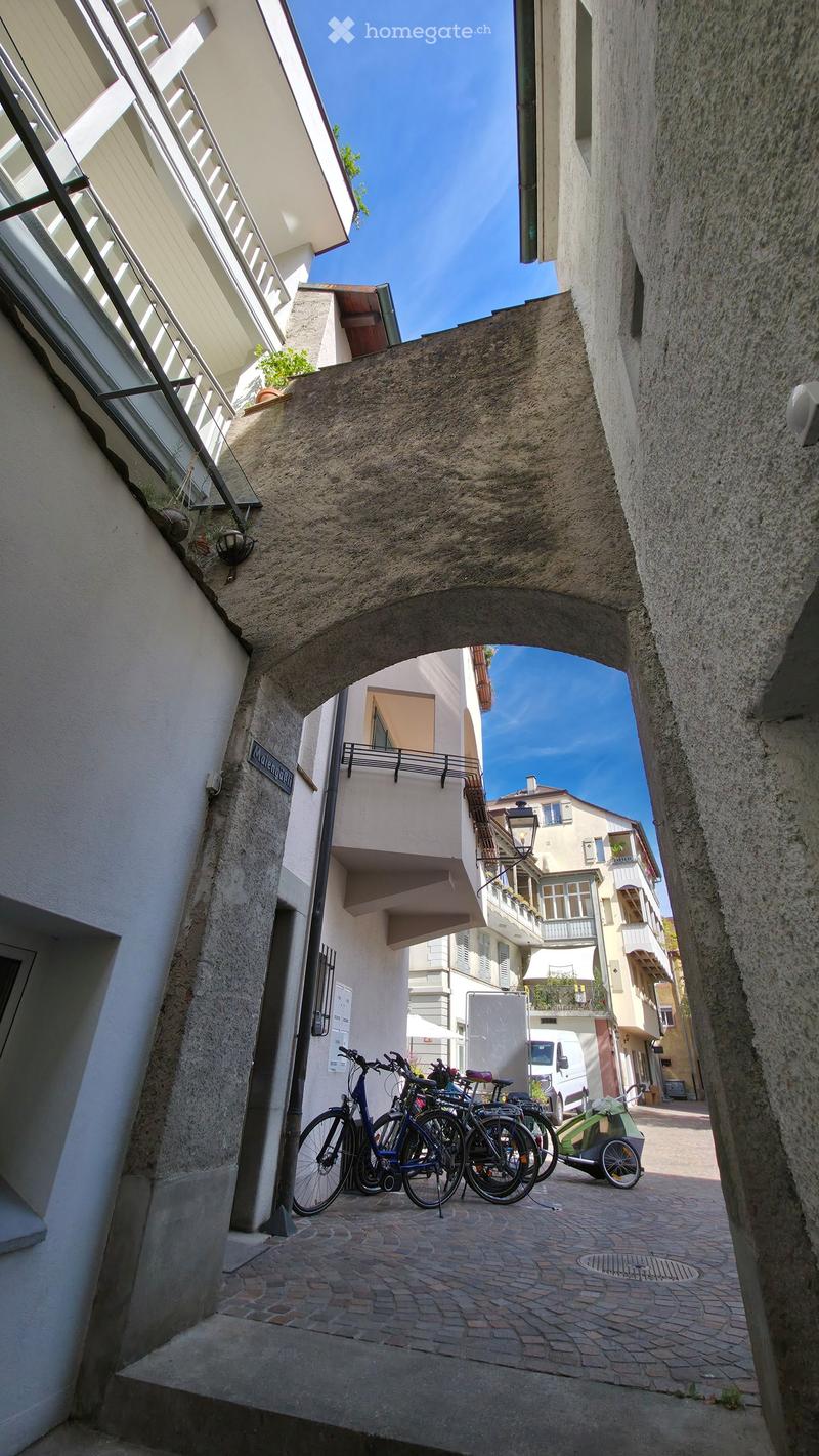A narrow, cobblestone alleyway flanked by tall buildings with balconies and windows, featuring bicycles parked along the left side.