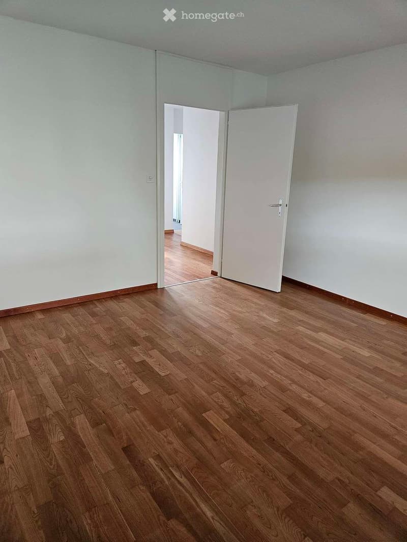 Empty room with wooden flooring and white walls, featuring a partially open white door leading to another room.