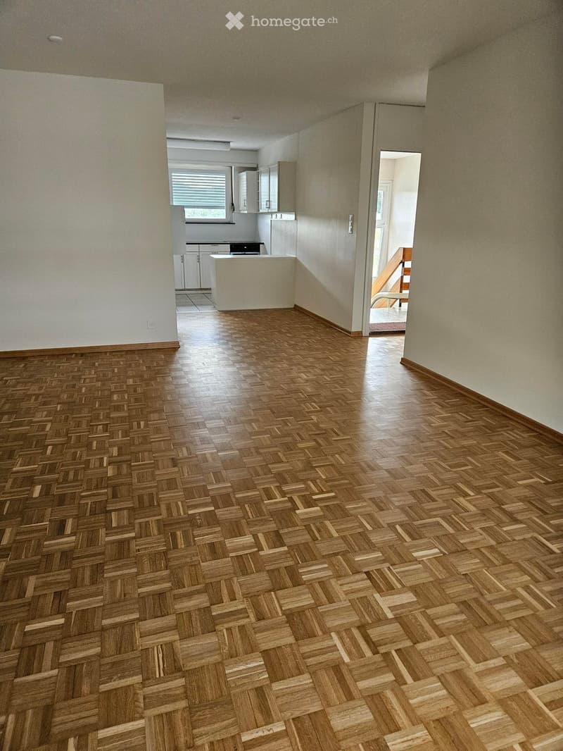 Empty room with wooden herringbone flooring, white walls, and a doorway leading to another room with cabinets and a window.