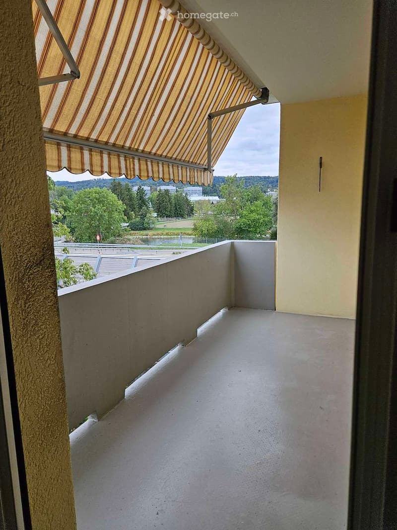Balcony with striped awning, concrete floor, and a view of a green landscape and buildings.