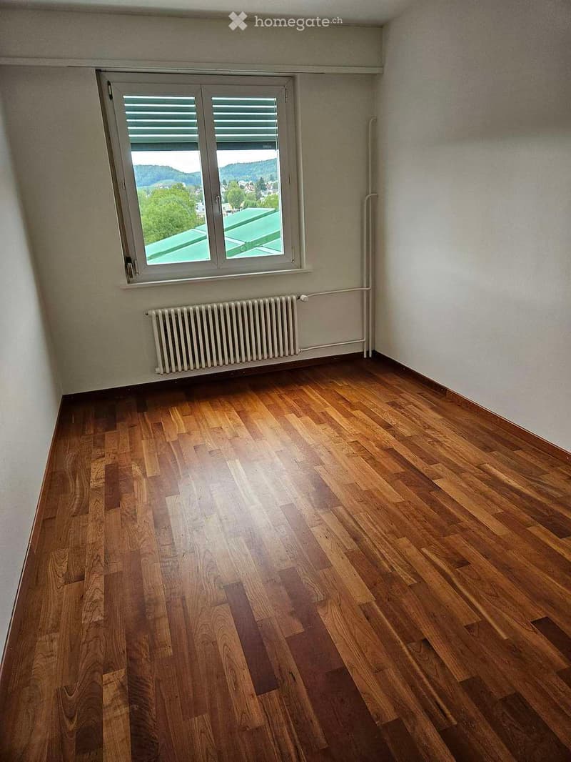 Empty room with wooden flooring, white walls, a window with blinds, and a radiator below the window.
