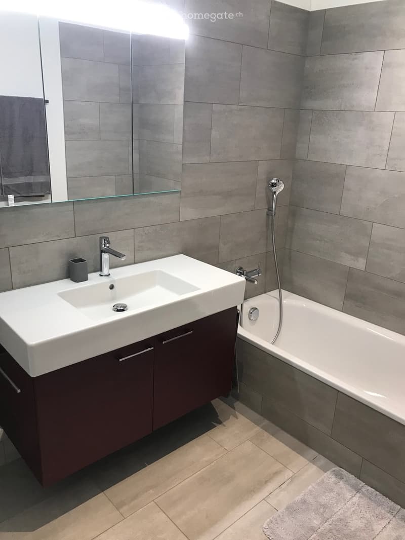 Modern bathroom with a double sink, dark wooden cabinet, bathtub, and large mirror. Grey tiles on walls and light wooden floor.