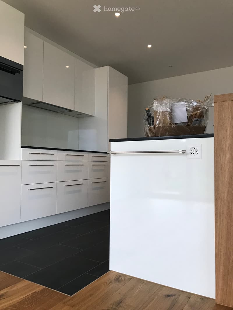 Modern kitchen with white cabinets, black countertop, and wooden flooring. An oven is visible on the left, and a small basket with items sits on the counter.