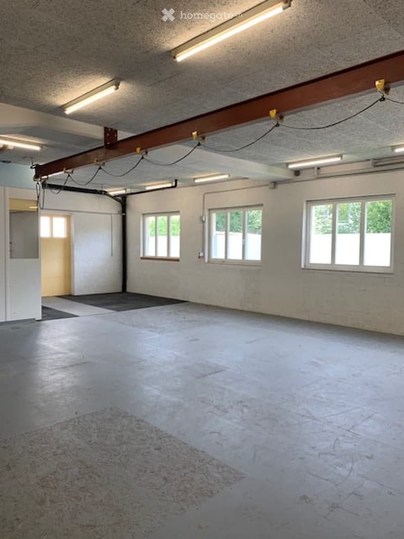 Empty industrial-style room with concrete floor, white walls, large windows, exposed ceiling beams, and fluorescent lighting.