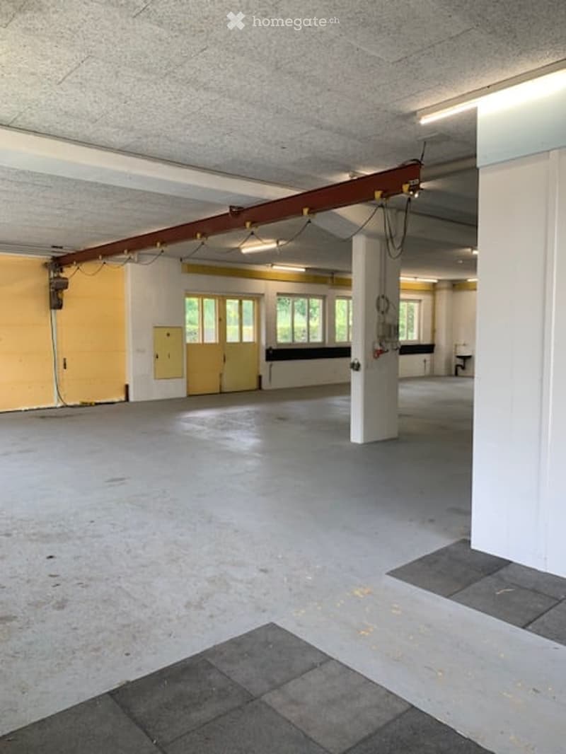Empty industrial-style room with concrete floors, exposed ceiling beams, and large windows. No furniture or equipment present.