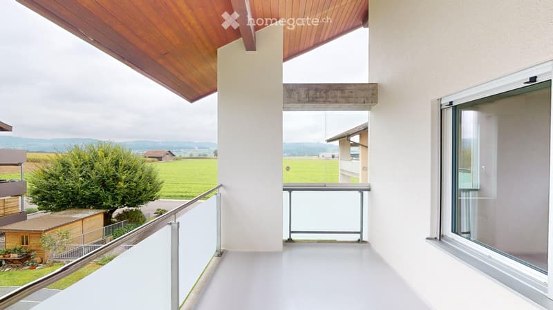 Modern balcony with wooden ceiling, white floor, glass railing, and scenic countryside view.