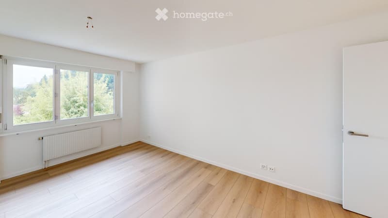 Empty room with white walls, wooden flooring, large windows, radiator, and a door.