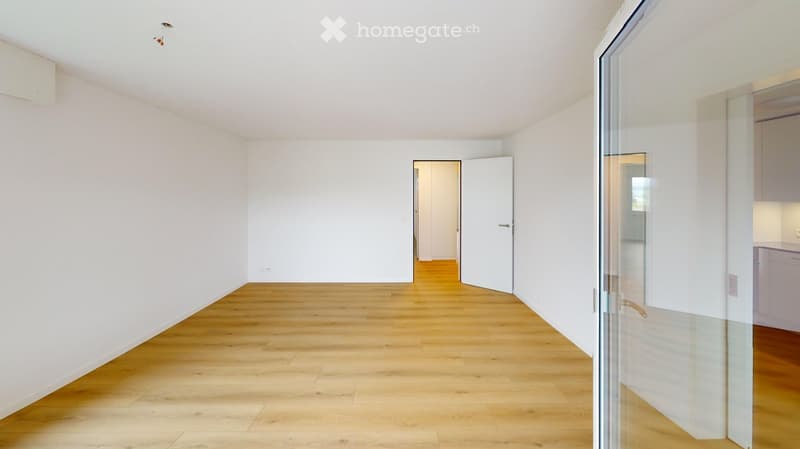 Empty room with light wooden flooring and white walls, featuring a glass door leading to another area.