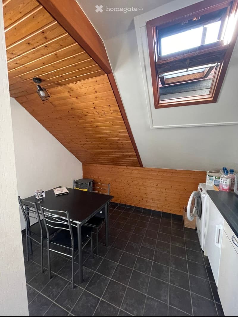Attic room with wooden ceiling, dark tiled floor, and a small dining area with a table and chairs. A washing machine is positioned near the corner, and a window with blinds allows natural light to enter.