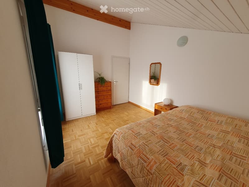 Bedroom with wooden flooring, a bed with patterned bedding, a white wardrobe, a wooden nightstand with a lamp, and a small plant on a dresser.