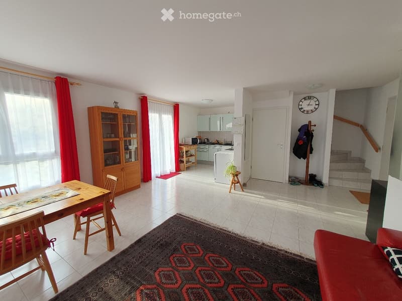 A well-lit living and dining area with a red sofa, wooden dining table with chairs, a wooden cabinet, a small kitchen area with a refrigerator, microwave, and a clock on the wall. There is also a staircase leading to another level.
